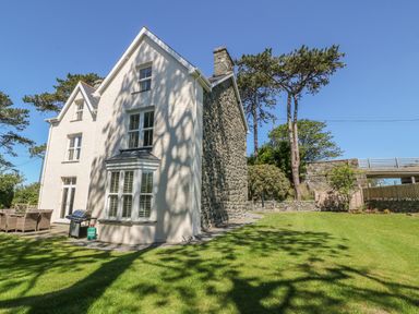 A two story house with white walls and stone side wall with a patio set and grill on a lawn at Tyddyn Goronwy in Talybont