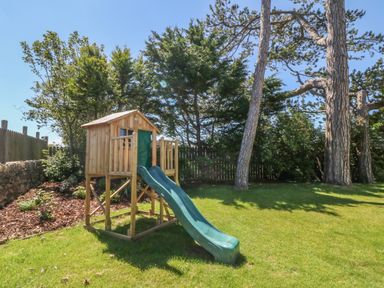 A wooden playhouse with a green slide in a fenced grassy yard with trees at Tyddyn Goronwy in Talybont