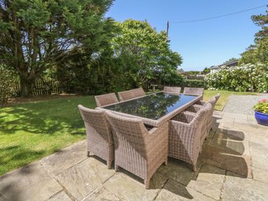 An outdoor patio area with a glass top table and eight wicker chairs surrounded by grass and trees at Tyddyn Goronwy in Talybont