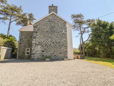 A stone house with a gravel driveway and outdoor seating area surrounded by trees at Tyddyn Goronwy in Talybont