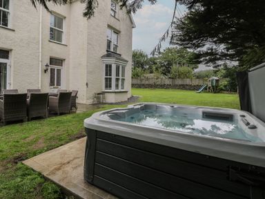 A backyard with a hot tub a dining table with chairs a playhouse with a slide and a white house at Tyddyn Goronwy in Talybont
