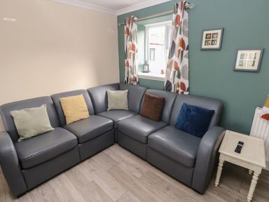 A corner sofa with cushions next to a window with patterned curtains and a small table with a remote control at Barn Cottage in Laugharne