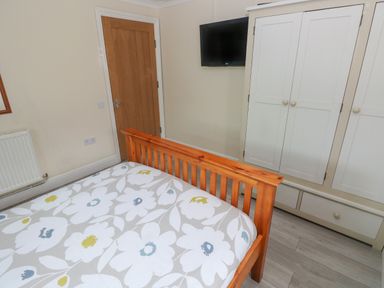 A bedroom with a wooden bed frame a floral duvet a wooden door a wall-mounted TV and a white wardrobe with drawers at Barn Cottage in Laugharne