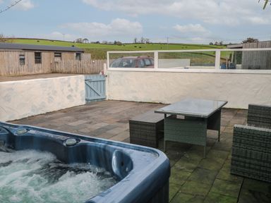 An outdoor patio with a hot tub a glass table with wicker chairs and a wooden fence at Barn Cottage in Laugharne