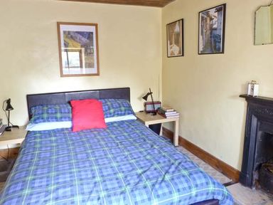 A bedroom with a bed blue checkered bedding and a red pillow two bedside tables with lamps framed pictures on the walls and a fireplace at Clooncorraun Cottage Ballinrobe County Mayo