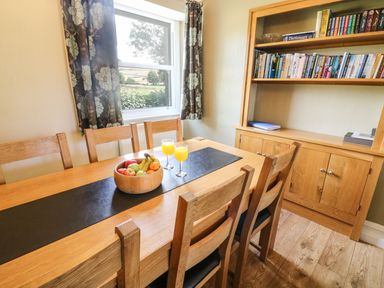 A dining room with wooden table and chairs a fruit bowl two glasses of orange juice a bookcase with books and window with curtains at Riverside View in Cark in Cartmel