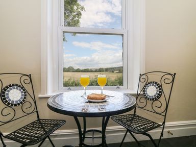 A small round tiled table with two glasses of orange juice and two black metal chairs by a window with a view of a field at Riverside View in Cark in Cartmel