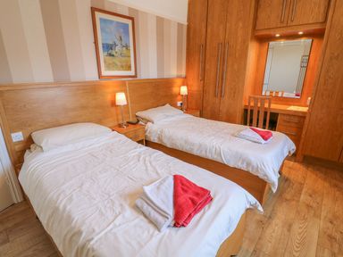 A bedroom with two single beds bedside tables lamps a painting and wooden wardrobes with a desk and chair at Riverside View in Cark in Cartmel