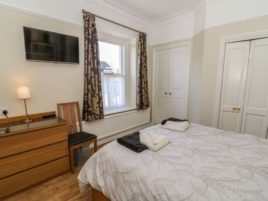 A bedroom with a bed two towels on it a wooden chair a chest of drawers a wall mounted TV and a window with curtains at Riverside View in Cark in Cartmel
