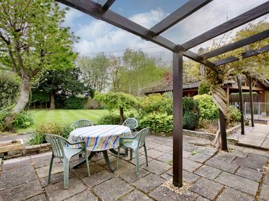 An outdoor patio with a table covered by a striped cloth and four plastic chairs next to a wooden pergola and garden at Ingleside Adsett Royal Forest of Dean