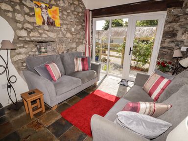 A sitting room with two grey sofas, a red rug, a wooden side table, stone walls, and glass doors at The Coach House in Wolfscastle