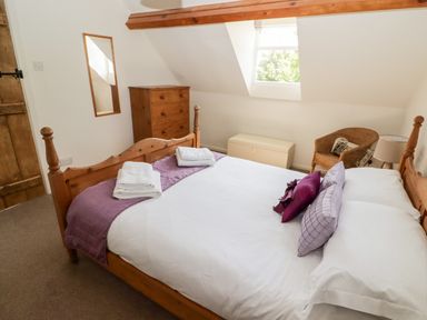 A bedroom with a wooden bed white bedding purple pillows a wooden chest of drawers a wicker chair and a window at The Coach House in Wolfscastle