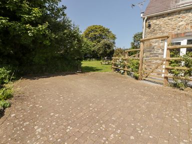 An outdoor paved area next to a stone building with wooden fence and gate leading to a grassy garden with trees at The Coach House in Wolfscastle