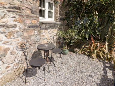 An outdoor seating area with a small round table and two metal chairs on a gravel surface next to a stone wall with a window and surrounding plants
