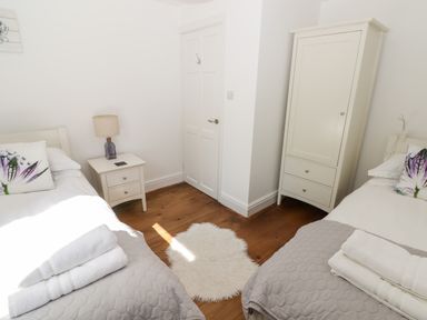 A bedroom with two single beds with grey covers and floral pillows a white nightstand with a lamp a white wardrobe a white door and a small white rug on wooden floor at The Duck House in Wolfscastle