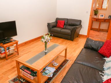 A living room with two black sofas a wooden coffee table with flowers a television on a wooden stand and a wooden floor at Mary Agnes Cottage in Cahirkeen near Allihies County Cork
