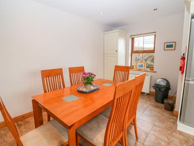 A dining area with a wooden table and six chairs a potted plant on the table a window with framed pictures a trash can and a cabinet in Mary Agnes Cottage in Cahirkeen near Allihies County Cork