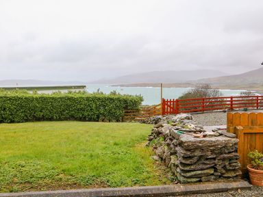 A garden with green grass a stone wall and wooden fences overlooking a body of water and mountains at Mary Agnes Cottage in Cahirkeen near Allihies County Cork