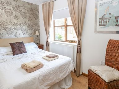 A bedroom with a double bed, a window with curtains, a bedside table with a lamp, and a wicker chair at The Haybarn in Necton near Swaffham