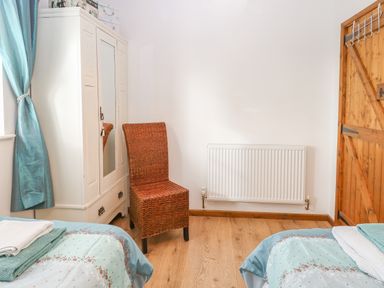 A bedroom with two beds, a white wardrobe with mirror, a wicker chair, a radiator, and wooden door at The Haybarn in Necton near Swaffham