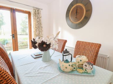 A dining table with a teapot set and flowers with wicker chairs and large wall clock at The Haybarn in Necton near Swaffham
