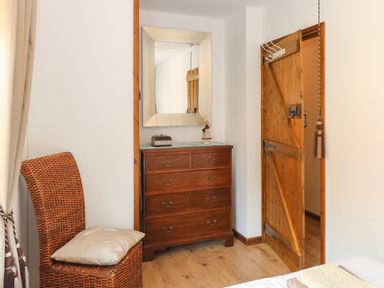 A corner of a room with a wicker chair with a cushion wooden chest of drawers a mirror and a wooden door at The Haybarn in Necton near Swaffham