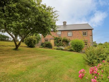 A brick house with multiple windows surrounded by a garden with trees bushes and flowers at Byrdir Cottage in Rhayader
