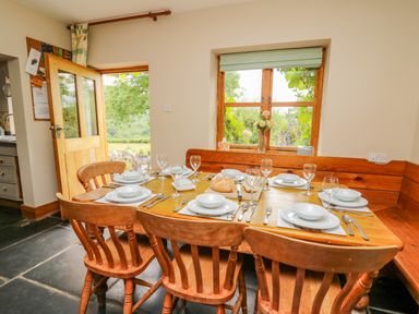 A dining area with a wooden table set for six with plates glasses and cutlery near a window and an open door at Byrdir Cottage in Rhayader