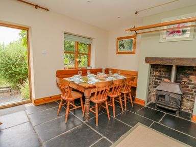 A dining area with a wooden table set for six and a wood burning stove in a corner at Byrdir Cottage in Rhayader
