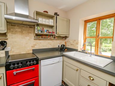 A kitchen with a red stove, dishwasher, sink by a window, cabinets and shelves with containers at Byrdir Cottage in Rhayader
