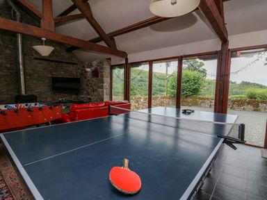 A game room with a ping pong table, foosball table, red sofas, a television, and large windows showing a stone wall and green hills outside at Byrdir Cottage in Rhayader