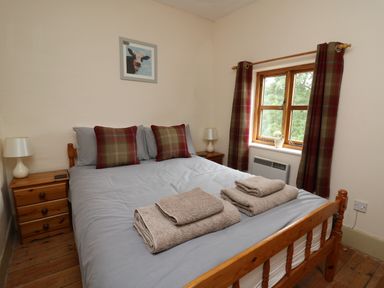 A bedroom with a wooden bed, two bedside tables with lamps, a window with plaid curtains, and folded towels on the bed at Byrdir Cottage in Rhayader