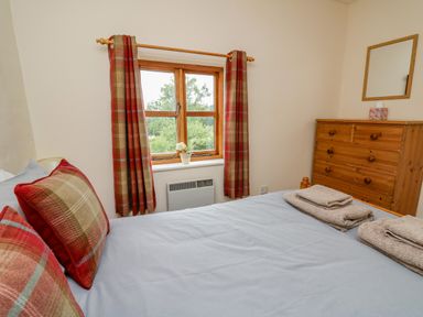 A bedroom with a bed, plaid pillows, folded towels, wooden dresser, mirror, window with plaid curtains and a small plant at Byrdir Cottage in Rhayader
