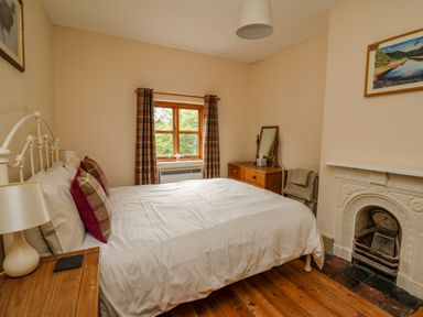 A bedroom with a bed pillows bedside table lamp window wooden dresser mirror and a fireplace at Byrdir Cottage in Rhayader