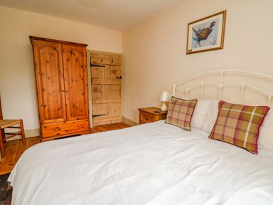 A bedroom with a wooden wardrobe a wooden door a bed with white bedding and plaid pillows and a nightstand with a lamp at Byrdir Cottage in Rhayader