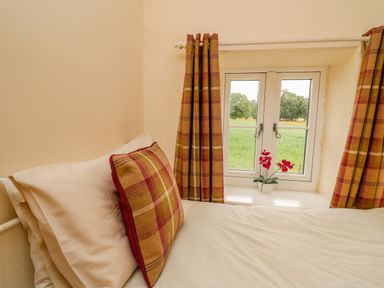 A bed with white sheets and a plaid pillow next to a window with plaid curtains and a small flower pot at Byrdir Cottage in Rhayader