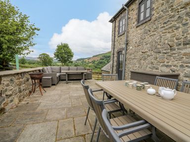 An outdoor patio with a dining table and chairs a stone wall and a sofa set at Byrdir Cottage in Rhayader