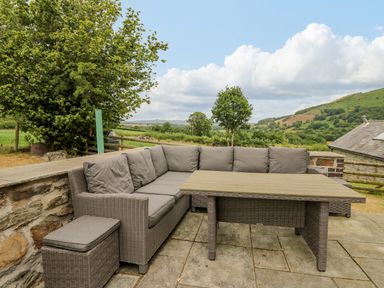 An outdoor patio with a corner sofa and table overlooking trees and hills at Byrdir Cottage in Rhayader
