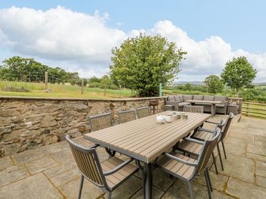 An outdoor patio with a dining table and chairs and a corner sofa set surrounded by trees and stone walls at Byrdir Cottage in Rhayader