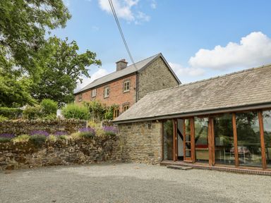 A stone and brick house with a slate roof next to a gravel driveway and stone wall with purple flowers at Byrdir Cottage in Rhayader