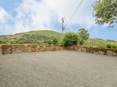 An outdoor gravel area bounded by a stone wall with trees and green hills in the background at Byrdir Cottage in Rhayader