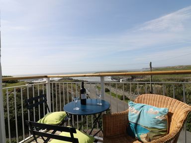 A balcony with a round table holding a bottle of wine and two wine glasses two chairs one with green cushions and one wicker chair with a cushion overlooking the sea at Seascape in Trearddur Bay