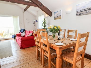 A dining area with a wooden table set for six beside a living room with a red sofa at Seascape in Trearddur Bay