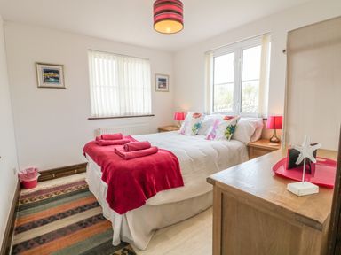 A bedroom with a double bed and red blankets two windows with blinds and wooden furniture with lamps at Seascape in Trearddur Bay
