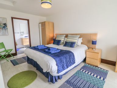 A bedroom with a double bed bedside tables rugs a green chair and an open door to a bathroom at Seascape in Trearddur Bay