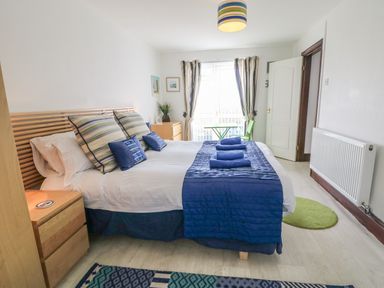 A bedroom with a double bed dressed in blue and striped pillows bedside tables and a small green table and chair near the window at Seascape in Trearddur Bay