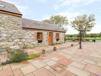 A stone cottage with a tiled patio and potted plants near trees and a lamp post at Poppy Cottage in Caeathro near Caernarfon
