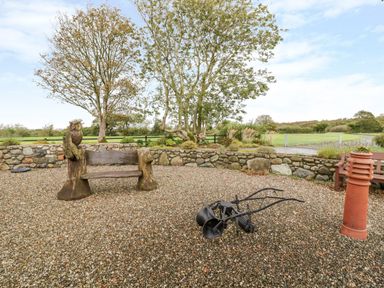 An outdoor gravel area with a carved wooden bench with owl decorations a metal garden plow a terracotta pipe and a stone wall with trees and fields beyond at Poppy Cottage in Caeathro near Caernarfon
