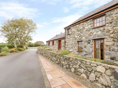 A stone cottage with wooden framed windows and a tiled walkway along the side of a paved road at Poppy Cottage in Caeathro near Caernarfon
