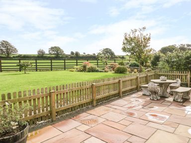 A fenced patio with stone table and benches overlooking a lawn and garden at Poppy Cottage in Caeathro near Caernarfon
